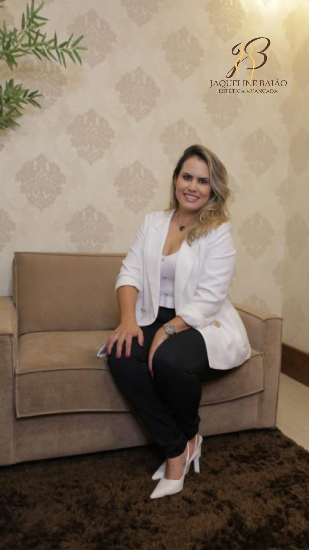 Dra. Jaqueline Baião sorrindo, sentada no sofá do consultório, entre a planta, a parede estampada e o tapete da sala